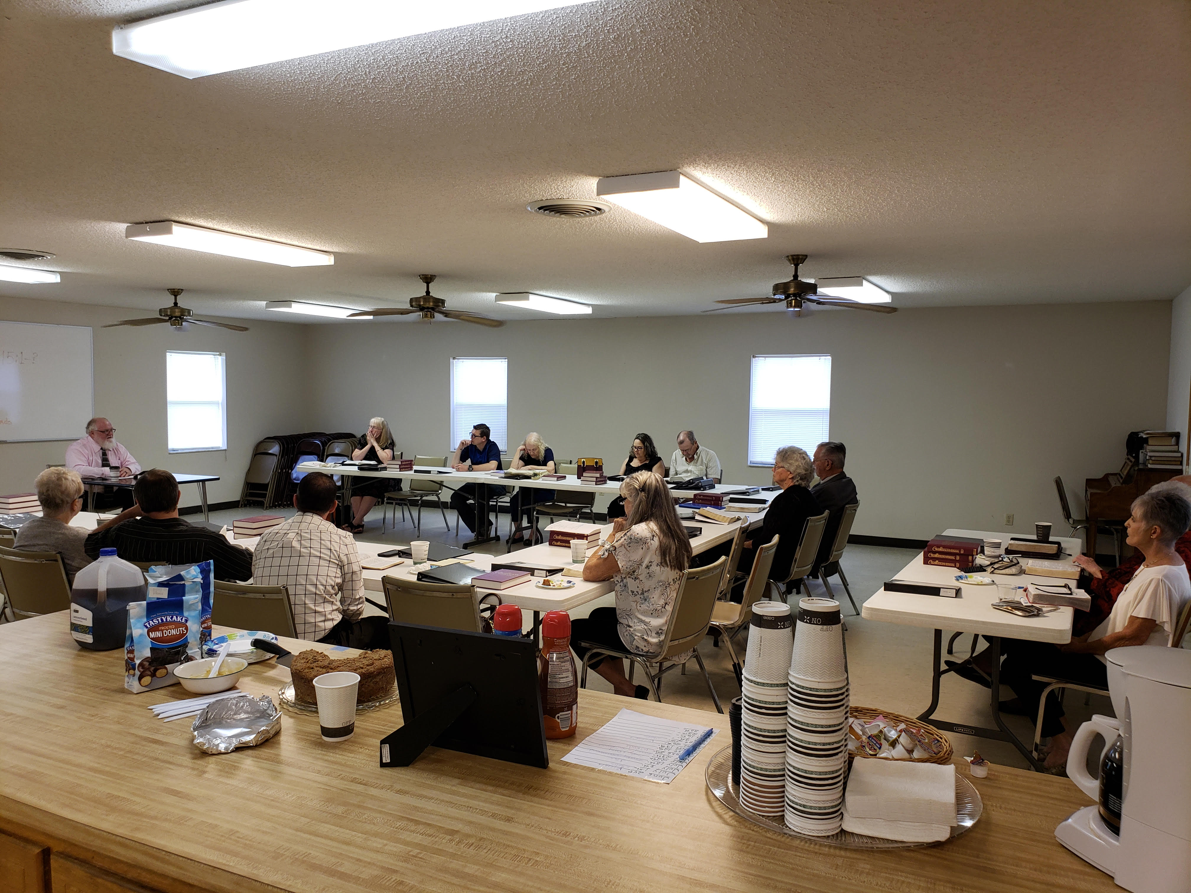 15 adults in semi-circle engaged in bible study with coffee and doughnuts